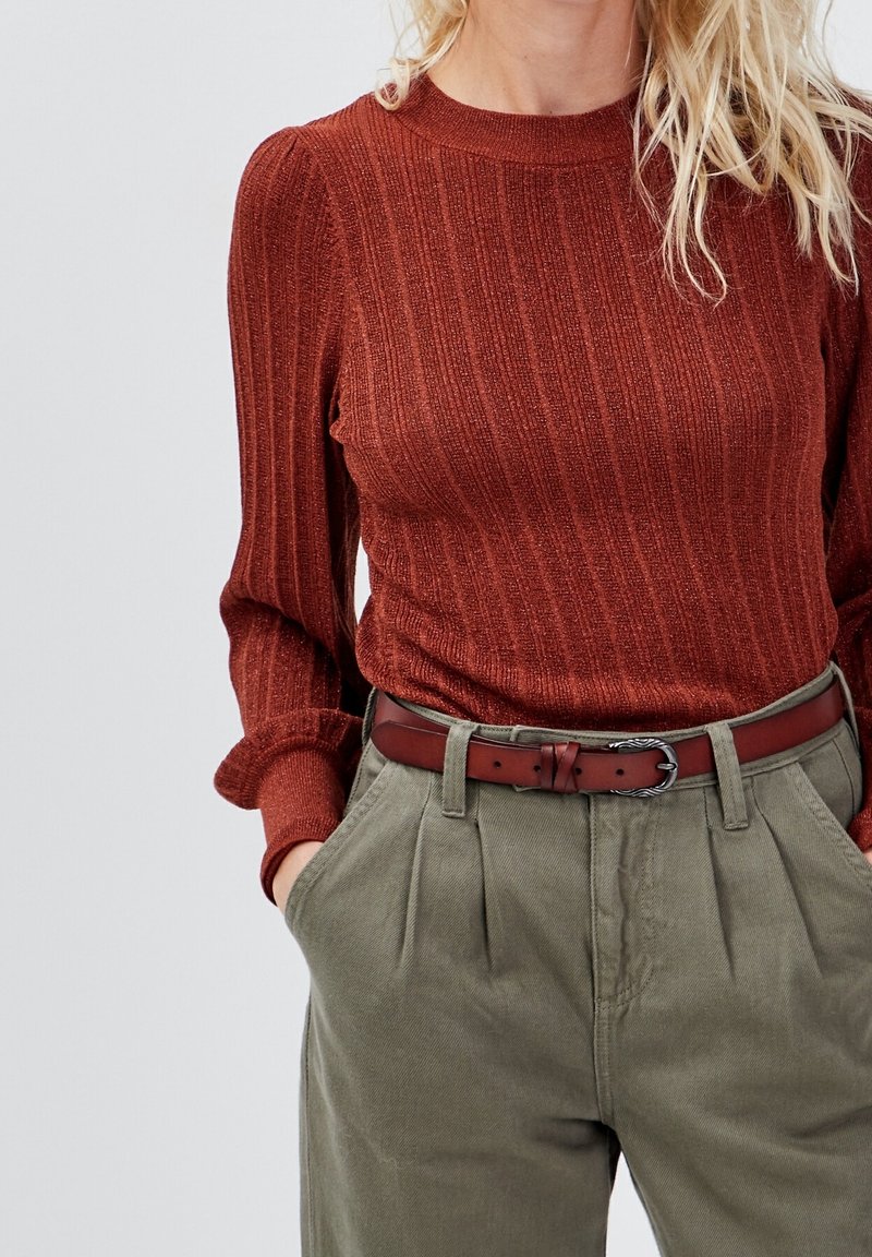 Femme portant un pull côtelé couleur rouille rentré dans un pantalon taille haute vert olive avec une ceinture en cuir rouge foncé.