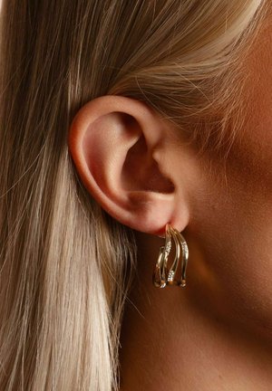 Goldene Creolen mit wellenförmigem Design und dezenten Diamantakzenten, getragen an einem Nahaufnahmeohr, die glatte Textur und polierte Oberfläche zeigen.