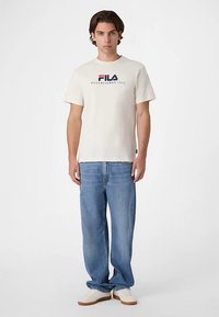 Kermanvärinen Fila-T-paita lyhyillä hihoilla, jossa on sininen ja punainen logo ja teksti. Yhdistetty vaaleansinisiin leveisiin farkkuihin ja valkoisiin lenkkareihin.