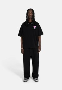 Crna oversized majica s ružičastim grafičkim motivom, uparena s crnim trenirkama. Model nosi srebrni nakit i sportske sive tenisice.