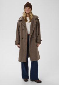 Palton din lână maronie cu rever mari, nasturi cu dublă închidere, buzunare laterale, purtat cu un top alb, blugi largi de culoare bleu închis și cizme maronii.