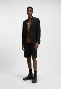 Blazer nero sopra una t-shirt grafica marrone, abbinato a pantaloni corti neri. Scarpe alte nere e calze con dettagli del logo. Sfondo semplice.