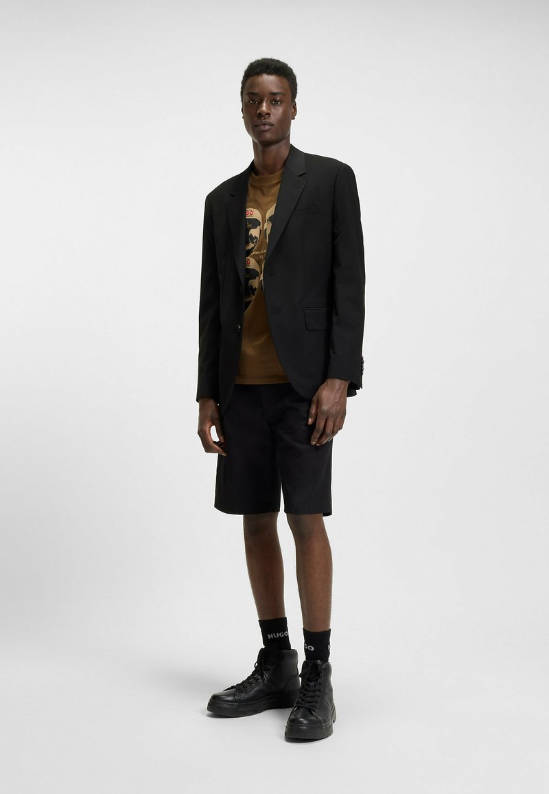 Blazer nero sopra una t-shirt grafica marrone, abbinato a pantaloni corti neri. Scarpe alte nere e calze con dettagli del logo. Sfondo semplice.