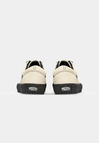 Zapatillas de ante beige con acentos en negro, forma redondeada, suela texturizada, costuras visibles y suela con marca. Vista trasera.