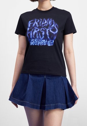 Woman wearing a black T-shirt with blue electric-style text and a pleated dark blue denim mini skirt against a plain background.