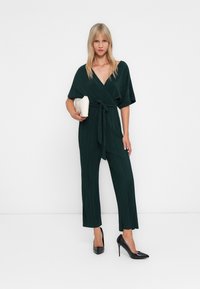 Kvinne iført en mørkegrønn plisserte jumpsuit med knytebelte, holder en kremfarget clutch og bruker svarte hæler foran en hvit bakgrunn.