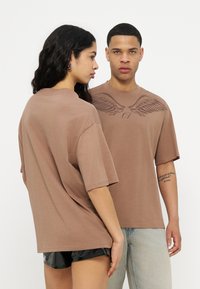 Dos personas llevan camisetas marrones holgadas, una de ellas de frente mostrando un diseño de alas en el pecho, la otra de perfil vistiendo pantalones cortos negros.