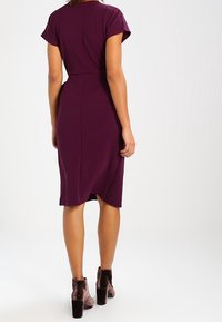 Donna che indossa un vestito prugna aderente fino al ginocchio con maniche corte e stivaletti in velluto fino alla caviglia, in piedi contro uno sfondo bianco semplice.