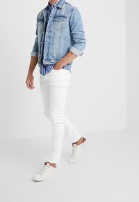 Chaqueta de mezclilla en azul claro, camisa a rayas en blanco y azul marino, pantalones slim-fit blancos y zapatillas blancas con detalles en marrón.