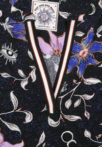 Svart tyg med färgglada blommönster, en ögon-design och en etikett. V-ringning med randig accent. Texturerad yta.