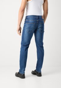 Mann iført blå jeans, svarte sko og en hvit ermeløs skjorte, stående mot en ensfarget hvit bakgrunn, bakfra.