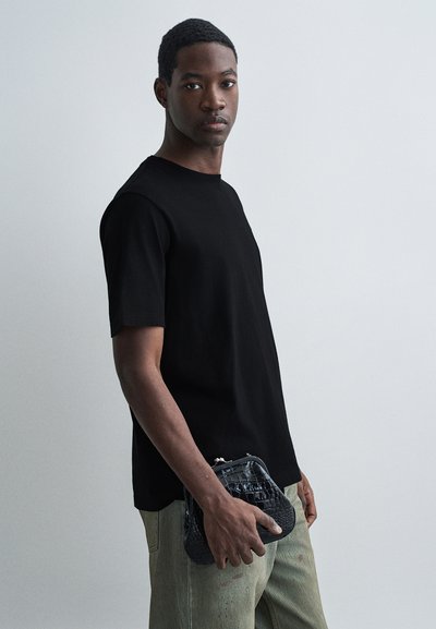Jeune homme portant un t-shirt noir et un pantalon clair, tenant une pochette noire texturée contre un fond uni clair.