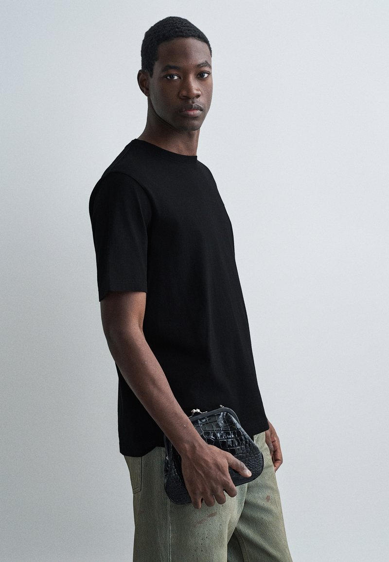 Joven con camiseta negra y pantalones claros sosteniendo un bolso de mano negro texturizado frente a un fondo claro.