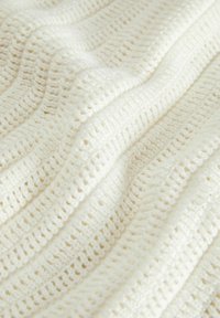 Cremefarbene Strickdecke mit einem strukturierten, wellenförmigen Muster und offenen Häkelstichen, die ein weiches und luftiges Aussehen verleihen.
