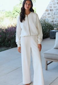 Woman standing outdoors wearing a white zip-up jacket and wide-leg pants, with greenery and patio furniture in the background.