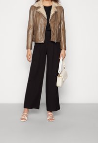 Giacca di pelle marrone con colletto peloso, abbinata a pantaloni neri a gamba larga e sandali bianchi con cinturini. Porta una borsa crema.