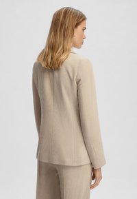 Blazer in corduroy beige con righe verticali, cuciture a punto singolo e colletto leggermente rialzato, indossato sopra pantaloni coordinati, visto da dietro.