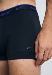 Schwarze Nike-Boxershorts mit einem lila Bund, der das Nike-Logo trägt. Der Stoff ist glatt und sitzt eng. Hand ruht auf dem Oberschenkel.