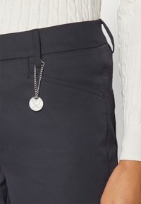 Pantaloni negri, croiți, dintr-un material neted, cu buzunare laterale și un detaliu dintr-un lanț cu pandantiv argintiu atașat de brâul pantalonilor.