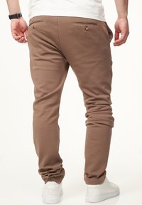 Brown sweatpants with a relaxed fit, featuring two back pockets with buttons, cuffed hems, and a soft texture. Paired with white sneakers.