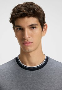 Gray short-sleeve shirt with a textured finish. Features a round navy collar with white trim accent. Model has medium curly hair.