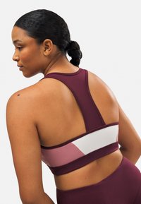 Sujetador deportivo burdeos con diseño de espalda nadadora, que presenta acentos en bloque de color en rosa claro y blanco. Textura suave, material elástico.