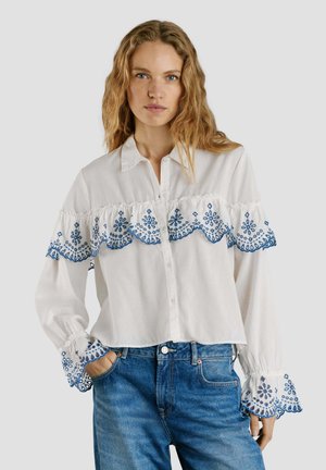 Frau trägt eine weiße Bluse mit blauer Blumenstickerei auf den Ärmeln und der Brust, kombiniert mit blauen Jeans, steht vor einem schlichten Hintergrund.