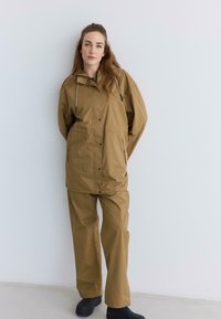 Femme portant une veste et un pantalon imperméables beige avec des bottes en caoutchouc noires, se tenant contre un mur blanc uni.