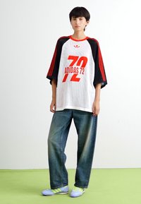 Camisa blanca de gran tamaño con acentos en rojo y negro, que presenta la impresión "ADIDAS 72". Combinada con unos pantalones vaqueros azul claro holgados y unas zapatillas deportivas azul claro.