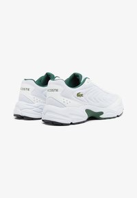 Vita atletiska skor med gröna accenter, texturerat meshmaterial, vadderad krage och gummisula. "LACOSTE"-logotypen på sidan.