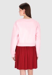 Sudadera rosa recortada con puños y dobladillo acanalados, combinada con una falda roja plisada, que muestra texturas suaves y colores vibrantes.