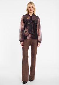 Blonde Frau in einer transparenten Bluse mit Blumenmuster und Rüschen sowie braunen, weiten Hosen, die vor einem weißen Hintergrund steht.