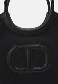 Černá taška typu tote s texturovaným látkovým tělem, kulatými koženými držadly a výrazným raženým logem na přední straně.