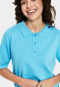 Svetlo modra polo majica s ovratnikom, kratkimi rokavi in tremi gumbi. Gladka tekstura in rebrast zaključek. Brez vzorcev ali dodatkov.