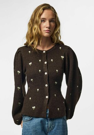 Femme aux cheveux blonds ondulés portant un pull marron foncé boutonné avec de petits motifs floraux blancs et un jean bleu sur un fond uni.