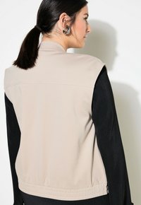 Beige ärmellose Weste mit geradem Schnitt, elastischem Bund, glatter Textur und schwarzem Rippstoff an den Seiten. Mit feinen Nähten akzentuiert.