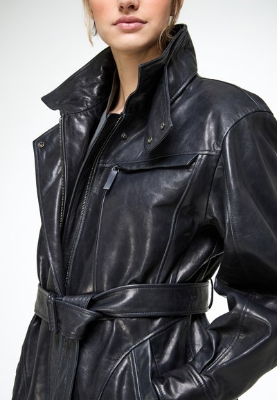 Vrouw met een zwarte leren jas met ceintuur, hoge kraag, ritssluiting aan de voorkant en een borstzak met rits, tegen een effen witte achtergrond.
