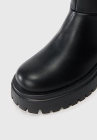 Bota de couro preta com ponta arredondada, sola com textura grossa preta e superfície lisa, apresentando um design elegante e sem detalhes visíveis.