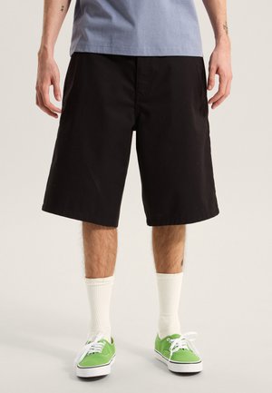 Pantaloncini neri in cotone con vestibilità rilassata, dotati di tasche laterali, abbinati a calzini bianchi fino al polpaccio e sneaker verdi. Design semplice e casual.