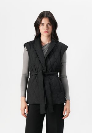 Svart quiltet vest med sjalkrage, tilpasset midje belte og ermeløs design. Bæres over en grå langermet topp.