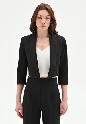 Donna con capelli castani fino alle spalle, indossa un blazer nero corto con maniche 3/4, una maglia bianca con scollo a V e pantaloni neri a vita alta, rivolta verso la fotocamera.