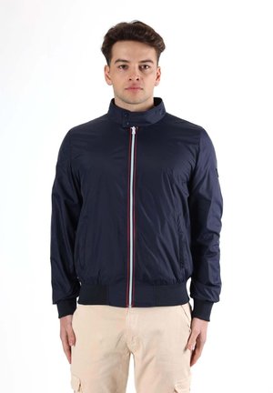 Un homme porte une veste bleu marine zippée à col montant et à fermeture éclair rayée, associée à un pantalon beige sur fond blanc.