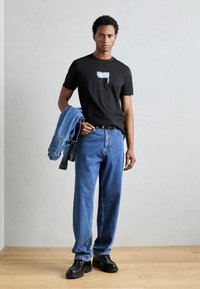 Camiseta negra con gráfico azul, jeans de denim azul holgados, chaqueta de denim colgada del brazo, botas negras, de pie sobre un suelo de madera.
