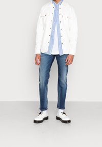 Hvid denimjakke over en blåstribet skjorte, kombineret med blå jeans og hvide sko med sorte såler og kontrasterende snørebånd.