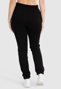 Schwarze Baumwoll-Sweatpants mit elastischem Bund, schmal zulaufenden Bündchen und Seitentaschen, getragen mit weißen Sportschuhen. Rückansicht des Kleidungsstücks.