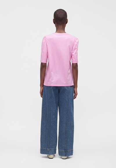 Personne debout de dos portant une chemise rose à manches courtes et un jean bleu à jambes larges avec des chaussures beiges sur un fond uni.