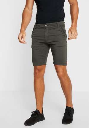 Bermudas verde oliva en una tela ligera, con bolsillos frontales, cierre de botón y un ajuste slim. Combinadas con zapatillas deportivas negras.