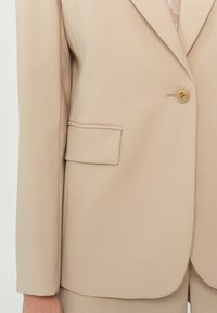 Blazer beige sur mesure avec fermeture à un bouton et poche à rabat sur le côté gauche, porté avec un pantalon assorti.