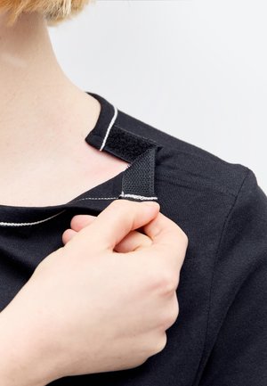 Hånd, der sikrer sort skjortes velcrolukning nær skulderen, viser let-åbent adaptivt tøjdétail på lys hud og blondt hår.