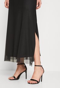 Black dress made of smooth fabric with a sheer hem and side slits, paired with black high-heeled sandals featuring ankle straps.